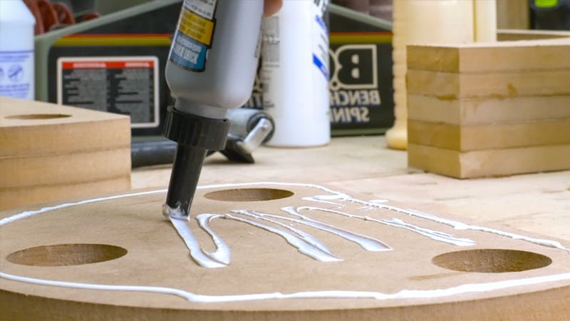 A shot looking down at a wooden mold where several thin, glue-covered wood strips are being carefully stacked. A glue roller is visible on the side