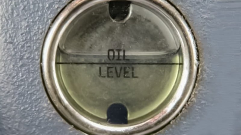 A technician's finger is pointing directly at the 'MAX' fill line on a circular oil sight glass of a vacuum pump. The clean, golden oil is filled perfectly to this line