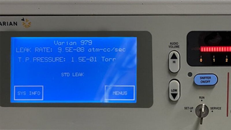 A macro shot focusing on the digital display of a helium leak detector. The screen is brightly lit, showing a graph and a specific numerical leak rate reading like 
