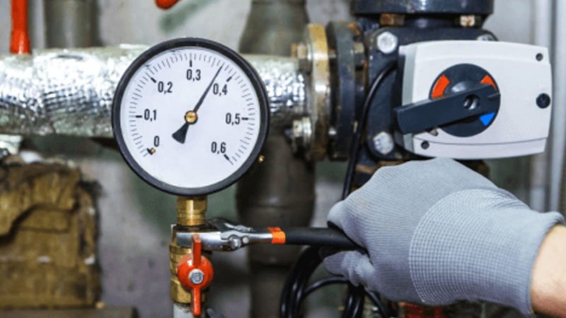 An industrial worker in overalls observes a large, analog pressure gauge with a red needle on a thick, heavy-duty pipe system.