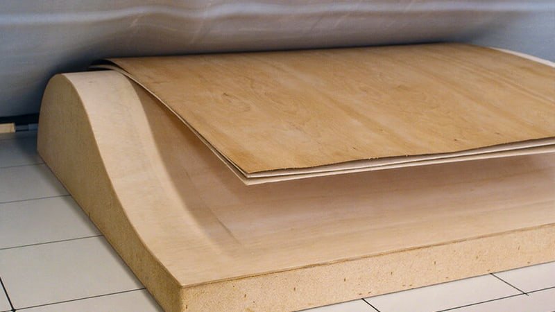 A woodworker in a sunlit workshop is pulling a freshly bent, laminated wooden chair back out of a large, clear vacuum press bag